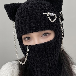 May include: A black crocheted balaclava featuring cat-ear shapes on top. The headgear is adorned with silver-toned metal chains and a safety pin holding white beads that spell out the letters W, E, and S in a horizontal arrangement.