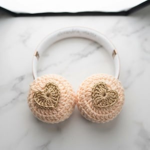 Könnte beinhalten: Weiße Kopfhörer mit hellpfirsichfarbenen Häkelbezügen auf den Ohrmuscheln. Jeder Bezug ist mit einem kleinen, mittig platzierten, metallisch goldenen Häkelherz verziert. Der Kopfbügel ist weiß mit metallisch goldenen Akzenten an den Gelenken.