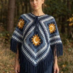 May include: A crochet poncho featuring a square pattern in shades of navy blue, light blue, and white. The design includes yellow sun motifs with white crescent moons at the center. The garment has a drawstring neckline and long navy blue fringe.