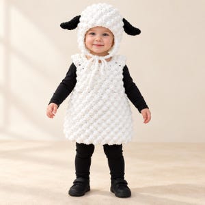 May include: A white crocheted sheep costume featuring a textured bobble stitch tunic and a matching bonnet with black ears. The outfit is worn over a black long-sleeved shirt and black leggings, paired with black shoes, creating a complete ensemble.