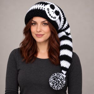 May include: A long, slouchy crochet beanie featuring a black and white striped pattern. The hat includes a large, circular white patch on the front with a black stitched skull face design and a black and white pom-pom at the end of the tail.