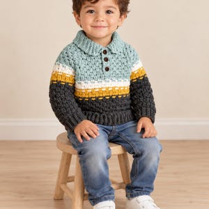 May include: A long-sleeve crochet polo shirt featuring a color-blocked design in light blue, white, mustard yellow, and dark gray. The shirt has a collared neckline with three dark brown buttons and a textured stitch pattern throughout the garment.