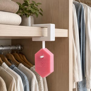 Puede incluir: Un gancho de plástico blanco montado en estante con un accesorio hexagonal de silicona rosa. El gancho blanco se sujeta a un estante de madera, mientras que el componente hexagonal rosa cuelga verticalmente con un icono de llama en relieve.