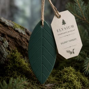 Può includere: Un ornamento sospeso a forma di foglia verde scuro con una venatura centrale e linee diagonali. Un cartellino di carta beige è attaccato con uno spago di juta, riportante il testo ELYSIUM, ODORIZANTE DE DULAP e SILENT FOREST, e un'icona di pino.