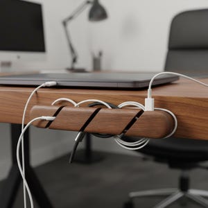 Puede incluir: Un organizador de cables de madera marrón oscuro montado en el borde de un escritorio. El bloque rectangular de madera cuenta con tres ranuras diagonales que sujetan cables de carga blancos y negros, manteniéndolos organizados para un portátil.