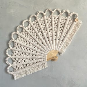 Puede incluir: Un abanico de mano con cordón de macramé color hueso anudado en un patrón geométrico, unido a un mango de madera clara. El borde superior tiene una serie de arcos en bucle y el borde inferior está rematado con un fleco de cordones sueltos.