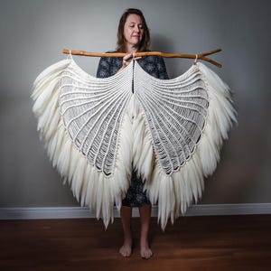 May include: A large, off-white macramé wall hanging shaped like a pair of expansive wings. The piece is constructed from thick, knotted cotton cord with long, soft, brushed fringe edges, suspended from a natural, horizontal wooden branch.
