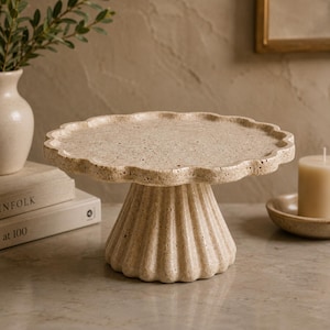 Puede incluir: Un soporte para pasteles de color beige con una textura porosa y moteada y un borde festoneado. El soporte presenta una base de pedestal ancha y estriada y una superficie superior plana y circular, hecha de un material similar a la piedra.