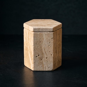 Op de afbeelding: Een zeshoekige container gemaakt van beige travertijnsteen, met een natuurlijke poreuze textuur en licht- tot donkerbruine aders. De houder is voorzien van een bijpassend, afneembaar zeshoekig deksel dat naadloos op de stenen behuizing rust.