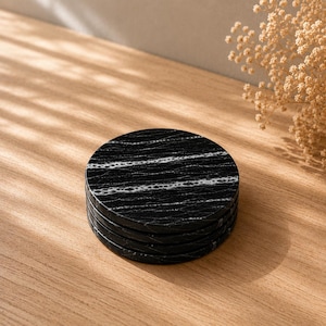 May include: A stack of four circular coasters made of black stone with prominent white veining patterns running horizontally across the surface. Each coaster has a smooth, polished finish and a consistent round shape, creating a uniform appearance.
