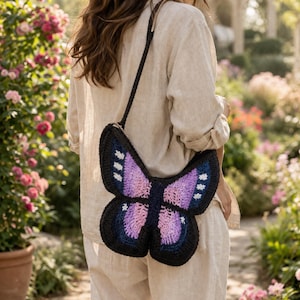 May include: A crochet shoulder bag shaped like a butterfly with a black border, purple and pink gradient wings, and small white accents. The bag features a long black strap and is worn over a light beige linen shirt and matching linen trousers.