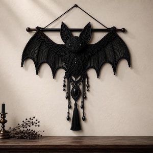 May include: A black macrame wall hanging shaped like a bat, suspended from a horizontal wooden rod with a cord. The bat features intricate knotted patterns, two pointed ears, and a central dangling element with black beads and a long tassel.