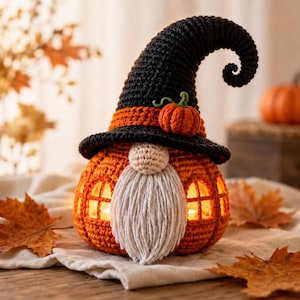 May include: A crocheted pumpkin-shaped gnome wearing a tall, curled black witch hat with an orange band and a small orange pumpkin accent. The gnome has a beige nose, a long white yarn beard, and orange crocheted walls with window cutouts.