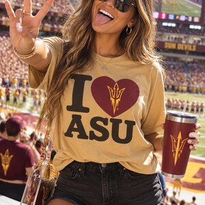 May include: A tan short-sleeved t-shirt featuring the black text 'I' and 'ASU' with a maroon heart containing a gold trident logo in the centre. The shirt is paired with black denim shorts and a maroon tumbler with a gold trident logo.