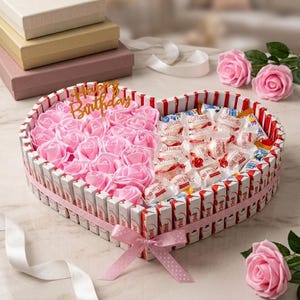 Könnte beinhalten: Eine herzförmige Geschenkbox aus Kinder-Schokoriegeln, gefüllt mit künstlichen rosa Rosen und weißen Raffaello-Kugeln. Ein goldfarbener Acryl-Aufstecker trägt die Aufschrift Happy Birthday. Das Arrangement ist mit einem rosa gepunkteten Band verziert.