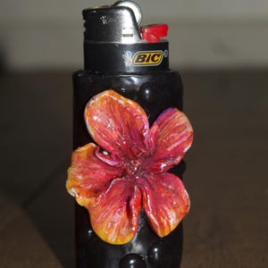 May include: A black Bic lighter featuring a three-dimensional, hand-sculpted flower in shades of red, orange, and yellow attached to the front. The lighter body is decorated with small, raised black circular accents surrounding the flower.