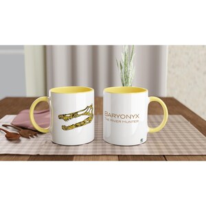 Könnte beinhalten: Zwei weiße Keramiktassen mit gelbem Innenraum und Henkel. Eine Tasse zeigt eine gelb-schwarze Grafik eines Dinosaurierschädels. Die andere Tasse zeigt den braunen Schriftzug BARYONYX THE RIVER HUNTER mit einem kleinen grünen Dinosaurier-Symbol.