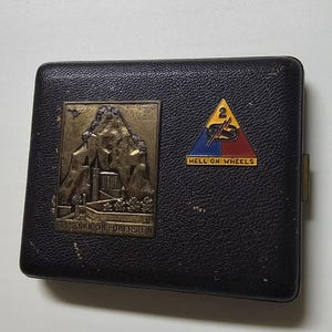 Puede incluir: Un estuche rectangular negro texturizado con dos emblemas metálicos. A la izquierda, un relieve en tono latón representa la Felsenkirche Oberstein. A la derecha, un parche triangular muestra el número 2, un rayo y el texto HELL ON WHEELS.