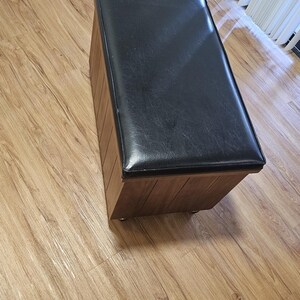 May include: A rectangular storage ottoman featuring a smooth black faux leather top cushion and a vertical wood-grain paneled base. The piece is supported by small, low-profile casters, providing a functional furniture item with a clean, structured design.