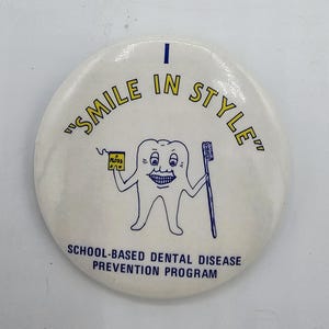 Könnte beinhalten: Ein runder weißer Ansteckbutton mit einer Cartoon-Zahnfigur, die eine gelbe Zahnseideschachtel und eine blaue Zahnbürste hält. Der Text lautet "SMILE IN STYLE" in Gelb und "SCHOOL-BASED DENTAL DISEASE PREVENTION PROGRAM" in Blau.