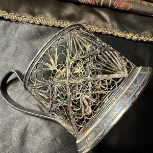 May include: An antique silver filigree teacup holder with a handle. The cup holder features intricate openwork patterns and a decorative rim. The metal has a tarnished appearance, suggesting age. The background includes a dark fabric with gold trim.