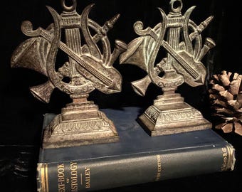 May include: Two antique bronze-colored bookends with musical instrument designs. Each bookend features a lyre, trumpet, and other instruments. The bookends are placed on a dark blue book.