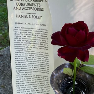 Puede incluir: Un libro titulado "The Complete Book of Garden Ornaments, Complements, and Accessories" de Daniel J. Foley. La portada del libro presenta un primer plano de una rosa roja oscura en un jarr&oacute;n de plata con tallas intrincadas. El texto de la portada describe el libro como una gu&iacute;a completa de los ornamentos de jard&iacute;n y sus usos.
