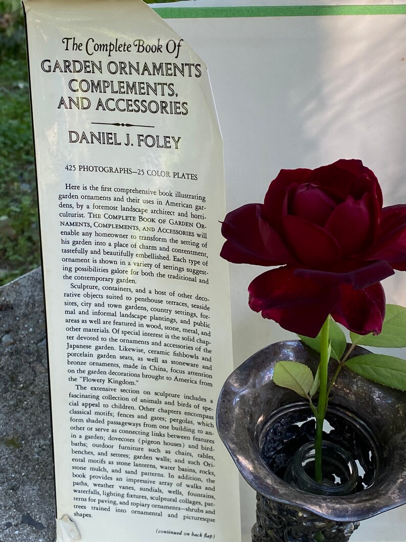 Puede incluir: Un libro titulado "The Complete Book of Garden Ornaments, Complements, and Accessories" de Daniel J. Foley. La portada del libro presenta un primer plano de una rosa roja oscura en un jarr&oacute;n de plata con tallas intrincadas. El texto de la portada describe el libro como una gu&iacute;a completa de los ornamentos de jard&iacute;n y sus usos.