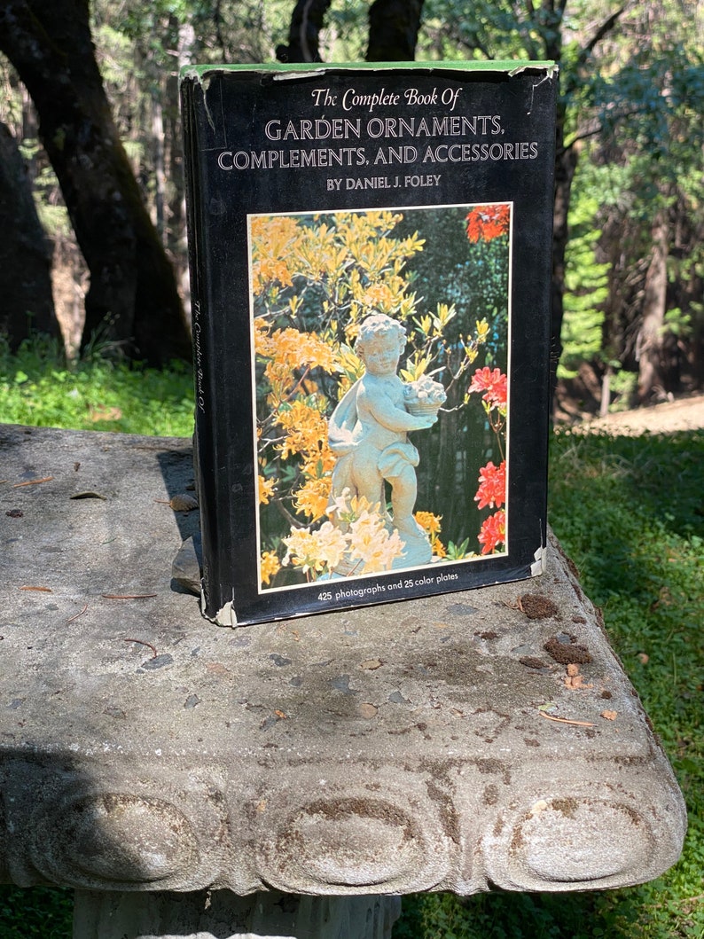 Puede incluir: Un libro de tapa dura negro titulado "The Complete Book Of GARDEN ORNAMENTS, COMPLEMENTS, AND ACCESSORIES" de Daniel J. Foley. La portada presenta una fotograf&iacute;a en color de una estatua de querub&iacute;n en un jard&iacute;n con flores amarillas y rojas.