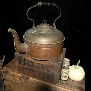 May include: A copper kettle with a wooden handle sits on a wooden crate with the words "Bon Air" printed on it. A small glass jar with a label that reads "Witch Hazel" and a white pumpkin are also on the crate.