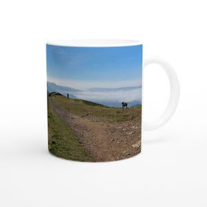 May include: A white ceramic mug featuring a photographic print of a grassy mountain trail under a clear blue sky. The image shows a dirt path winding through green hills, a distant figure, a small dog, and a valley filled with low-lying clouds.