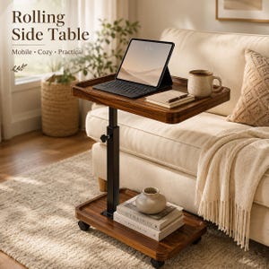 May include: A rolling side table with a dark wood finish, featuring a height-adjustable black metal post and four caster wheels. The top surface holds a tablet with a keyboard, a notebook, and a mug. A base shelf holds two stacked books and a vase.