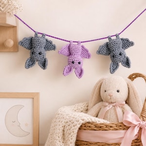 Puede incluir: Una guirnalda de ganchillo con dos murciélagos grises y uno central de color lavanda colgados de un hilo morado. Debajo, un conejo de peluche beige se sienta en una cesta tejida con una cinta de color rosa claro.