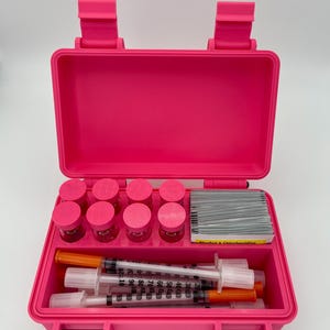 May include: A bright pink rectangular storage case containing eight small glass vials with pink caps, a stack of rectangular alcohol and chlorhexidine prep pads, and several clear plastic syringes with orange caps arranged in a lower compartment.