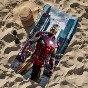 Può includere: Un telo mare rettangolare con una stampa grafica di Iron Man in un'armatura rossa e oro con un reattore arc luminoso sul petto, accompagnato da un cappello di paglia beige e occhiali da sole con montatura nera su una superficie sabbiosa.