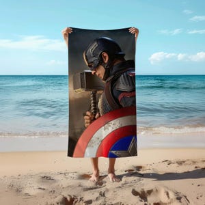 Op de afbeelding: Een rechthoekige strandhanddoek met een grafische print van een personage in een donkerblauw tactisch helm en pak, die een grote zilveren hamer vasthoudt achter een rond rood, wit en blauw schild.