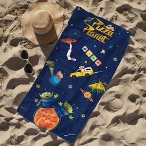 Może przedstawiać: Ciemnoniebieski ręcznik plażowy z logo Pizza Planet, żółtą ciężarówką dostawczą, zielonymi trzyokimi kosmitami, kawałkiem pizzy pepperoni i napisem OOOO. Ręcznik jest pokazany obok słomkowego kapelusza w kolorze piaskowym i czarnych okularów.