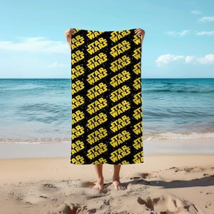 May include: A rectangular beach towel featuring a repeating pattern of the yellow Star Wars logo set against a black background with small white dots resembling stars. The towel is held upright, displaying the full graphic design clearly.