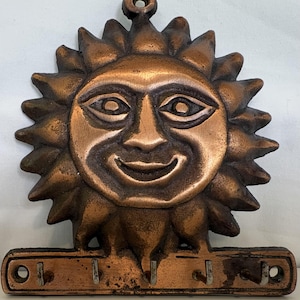 May include: A copper-toned metal wall-mounted key holder featuring a central sun face design with radiating triangular rays. The base includes five small hooks for hanging items and a circular loop at the top for mounting to a wall surface.