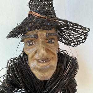May include: A handcrafted figurine featuring a sculpted face with detailed facial features, including painted eyes and eyebrows. The figure wears a wide-brimmed hat and a collar constructed from dark, woven natural fibers with a light brown accent.