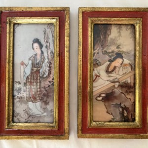 May include: A pair of rectangular wall art pieces featuring hand-painted scenes of figures in traditional attire. Each piece is encased in a deep red wooden frame with gold-painted inner trim, showcasing detailed illustrations of people.