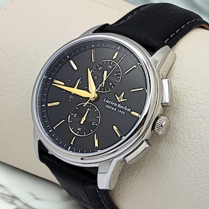Könnte beinhalten: Eine silberfarbene Chronographen-Armbanduhr mit dunkelgrauem Zifferblatt, goldfarbenen Zeigern und Stundenmarkierungen sowie einem schwarzen Lederarmband. Das Zifferblatt zeigt den Text Lucien Rochat DEPUIS 1925, zwei Hilfszifferblätter und ein Datum.
