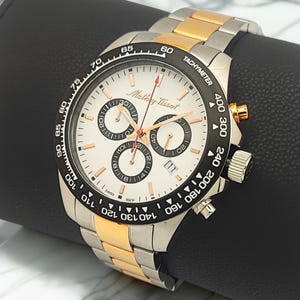 Può includere: Un orologio cronografo Mathey-Tissot con quadrante bianco, tre sotto-quadranti neri e datario. L'orologio presenta una lunetta tachimetrica nera e un bracciale in acciaio inossidabile bicolore con maglie color argento e oro.