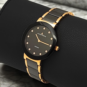 May include: A Jacques Lemans wristwatch featuring a round black dial with gold-tone hands and crystal hour markers. The watch has a black ceramic bezel and a link bracelet that combines black ceramic centre links with polished gold-tone metal edges.