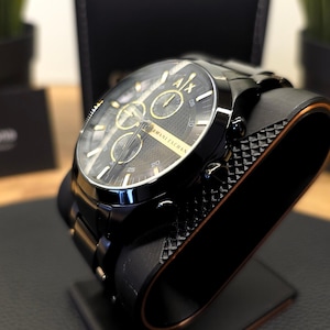 May include: A black Armani Exchange chronograph watch with a round face, gold-tone hands, and gold-tone hour markers. The dial features three sub-dials, the A|X logo at the twelve o'clock position, and the text Armani Exchange printed in gold.
