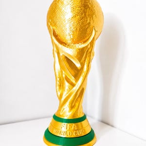 Peut inclure: Un trophée de couleur dorée avec un sommet sphérique soutenu par une base texturée et torsadée. La base comprend une bande verte avec le texte en relief FIFA WORLD CUP. L'objet présente une finition métallique et une structure verticale.