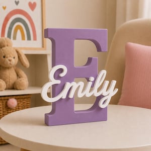 Può includere: Una grande lettera E in legno color viola opaco con il nome Emily scritto in corsivo bianco sulla parte anteriore. La lettera è autoportante e presenta una finitura liscia e solida, fungendo da elemento decorativo per scaffali o tavoli.