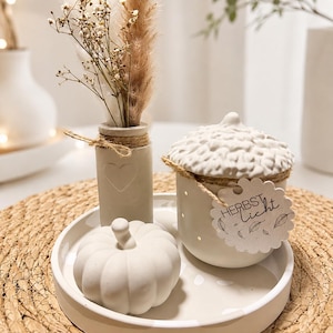 Peut inclure: Un plateau en céramique blanche contenant un petit vase avec des herbes séchées, une figurine de citrouille blanche et un récipient en forme de gland blanc. Le récipient a un couvercle texturé et une étiquette en papier avec HERBSTlicht.