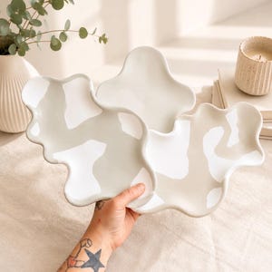 Peut inclure: Trois assiettes en céramique aux bords irréguliers et ondulés avec une finition lisse et brillante. Les assiettes présentent un motif organique abstrait alternant des tons blanc crème et beige doux, créant un aspect fluide et nuageux.