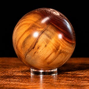 Peut inclure: Une sphère en bois poli présentant un motif de grain naturel avec des tourbillons de tons beige clair, brun doré et brun foncé. La sphère est soutenue par un petit support circulaire en acrylique transparent, affichant une surface lisse.